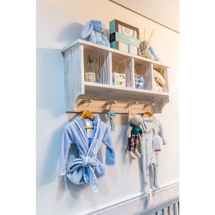 Distressed white Orchid wall shelf with four cubbies and three hooks, styled with baby clothes and toys, ideal for nursery storage
